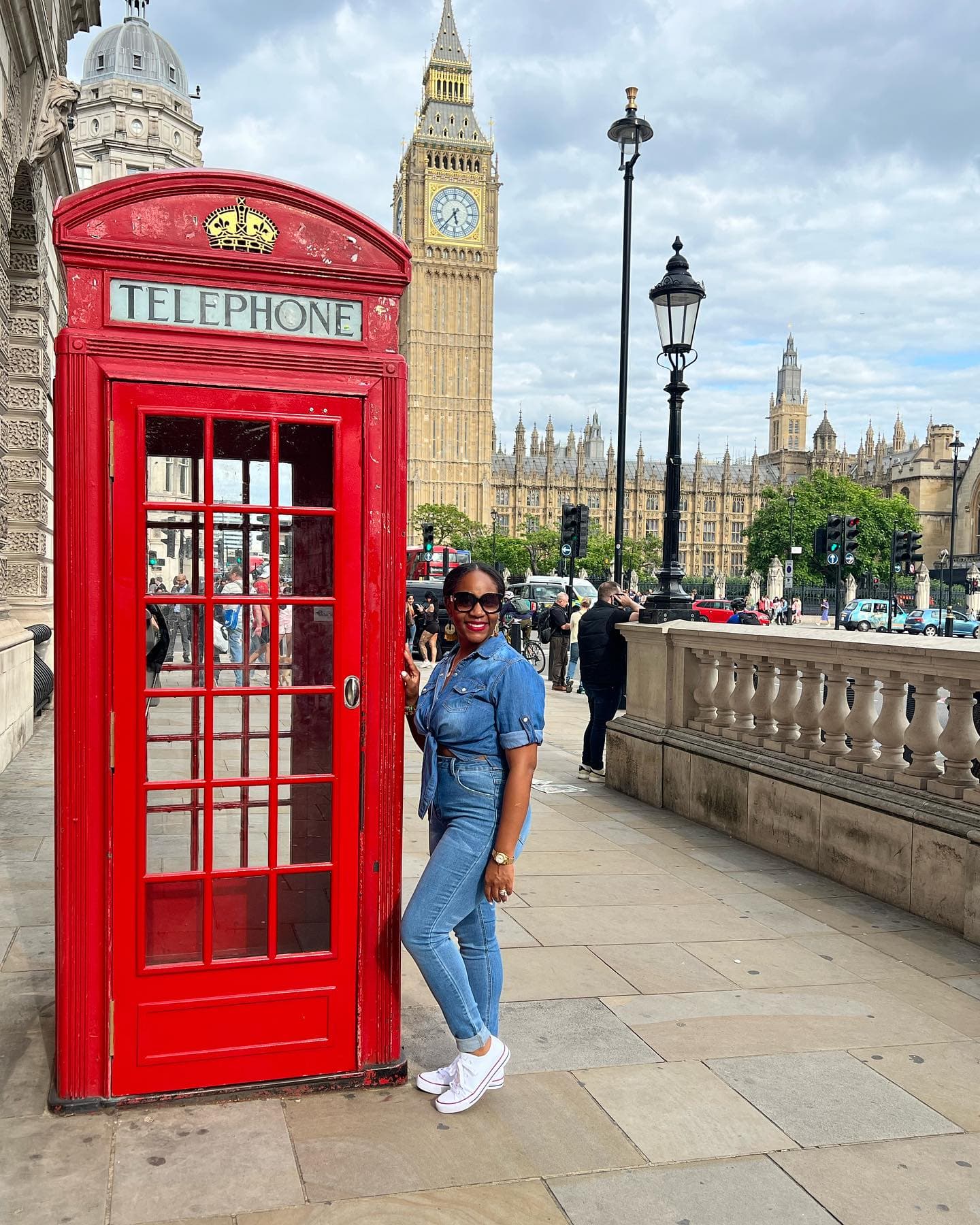 Neké Akua in a knotted denim shirt leaning against a red telephone box, with Big Ben behind her.
