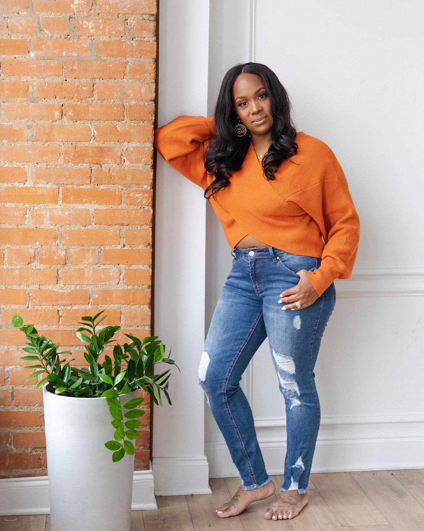 Neké Akua barefoot in an orange crossover knit and distressed jeans, leaning against a white wall next to exposed brick and a tall ZZ plant.