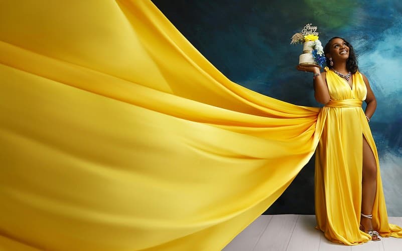 Neké Akua in a sweeping yellow satin gown holding a two-tier birthday cake trimmed with blue and yellow blooms.