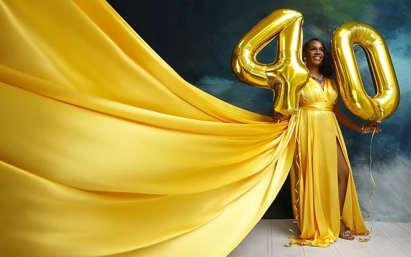 Neké Akua in a yellow satin gown holding gold foil balloons, the dress's long train sweeping the length of the studio floor.