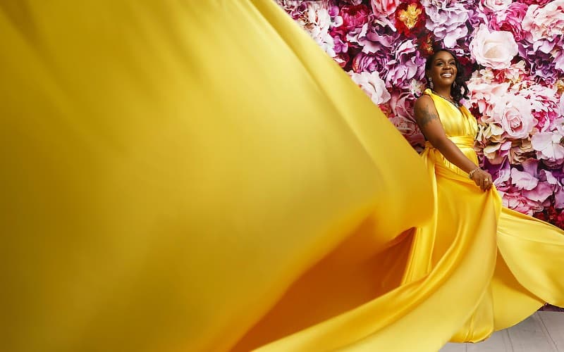 Neké Akua laughing in a yellow satin gown against a wall of pink and blush flowers, her train caught mid-motion across the frame.