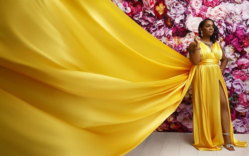 Neké Akua in a yellow satin gown with a high slit and silver heels, standing before a floor-to-ceiling pink flower wall with her train spread wide.