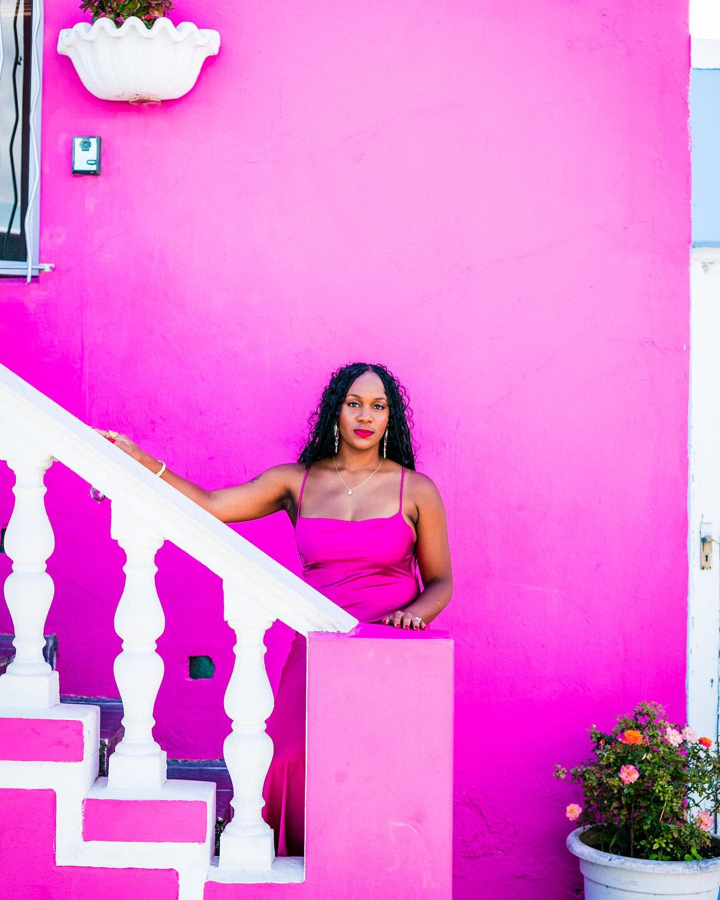 Neké Akua in a pink dress beside a white staircase against a magenta Bo-Kaap wall.