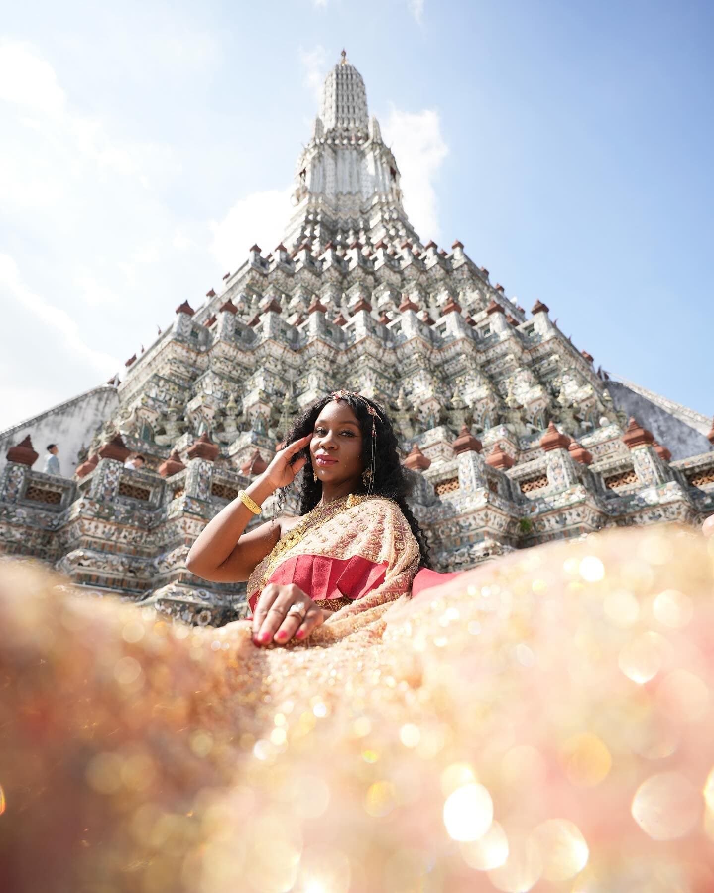 Neké Akua in a blush and gold traditional Thai costume, seated at the foot of the mosaic-tiled spire of Wat Arun in Bangkok.