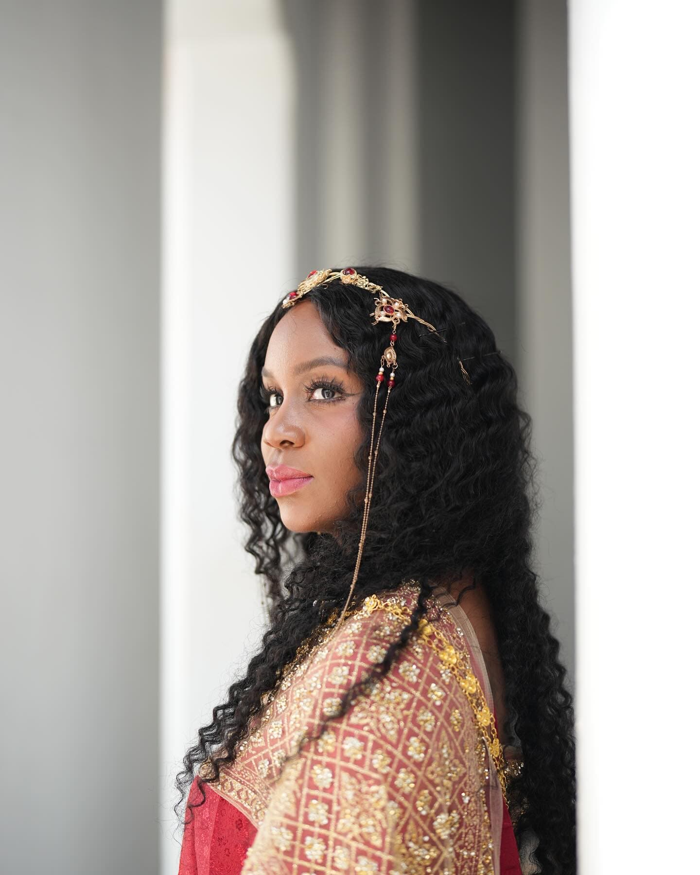 Close portrait of Neké Akua in a red and gold embroidered outfit and a jewelled gold headpiece, looking back over her shoulder against a soft grey stone backdrop.