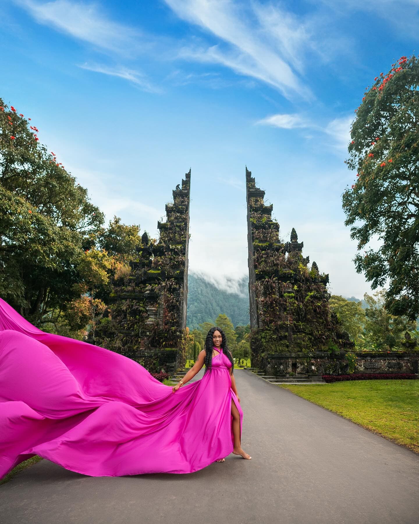 Neké Akua holding out the train of her magenta gown in front of a Balinese split gate beneath a wide blue sky.