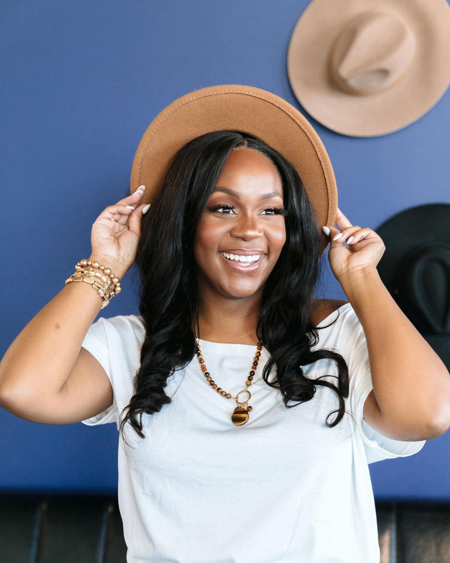 Neké Akua laughing as she sets a tan felt hat on her head, wearing a white tee and gold bracelets against a deep blue wall hung with more hats.