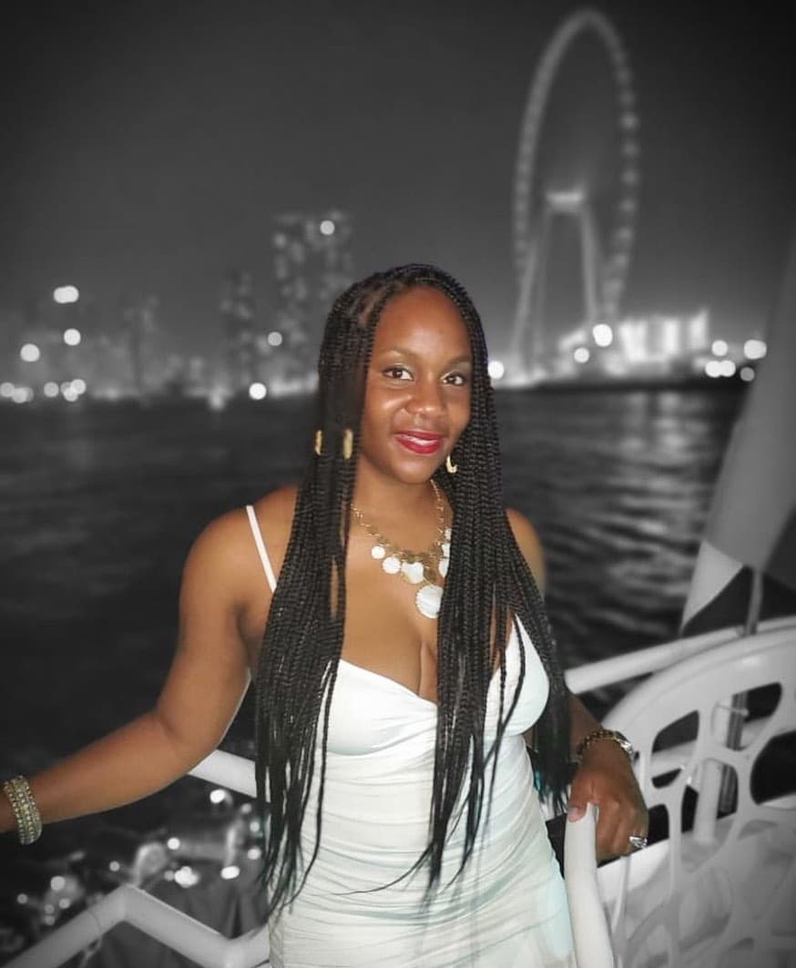 Neké Akua in a white dress and long braids, leaning on a boat railing at night with a lit-up waterfront skyline and a giant observation wheel behind her.