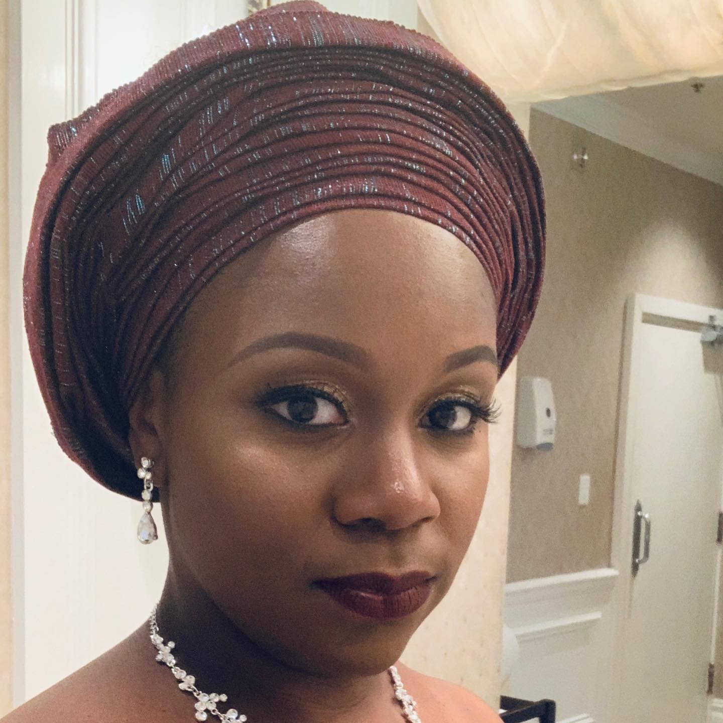 Neké Akua in a rose-brown metallic gele head wrap with crystal drop earrings and deep berry lipstick.