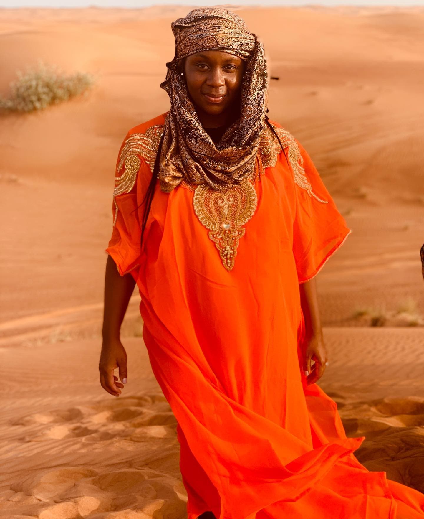 Neké Akua on the dunes at golden hour in an orange kaftan with gold beadwork and a matching patterned headscarf.