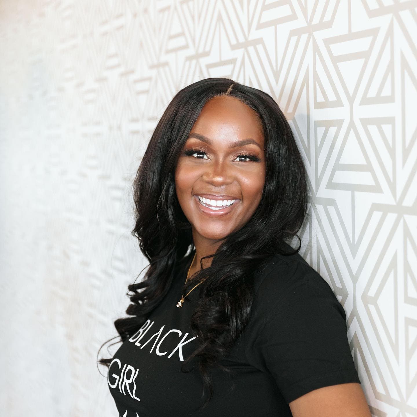 Neké Akua laughing in a black graphic tee against a beige geometric-print wall.