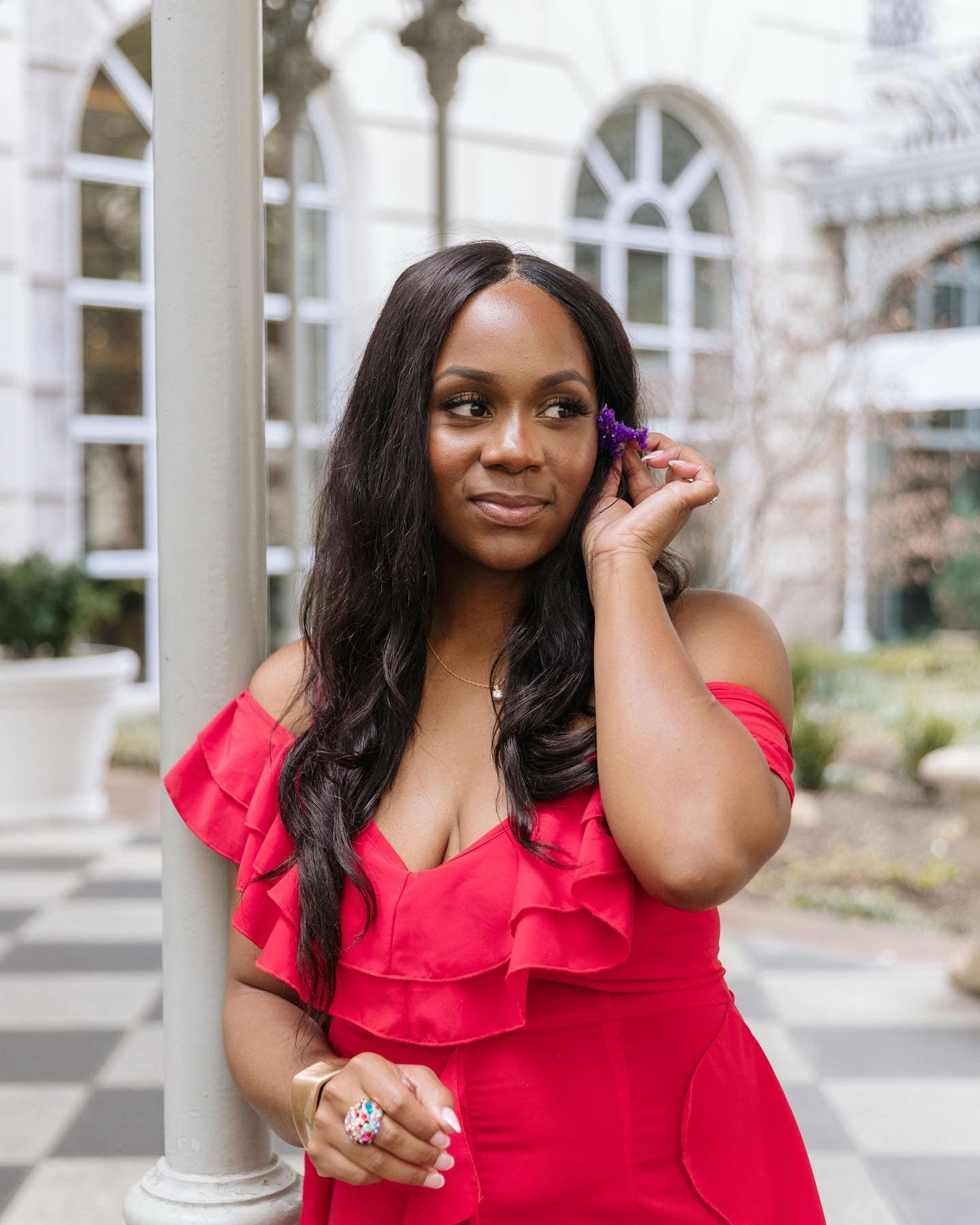 Neké Akua in a red ruffled off-the-shoulder dress on a checkerboard terrace, tucking a purple bloom behind her ear.