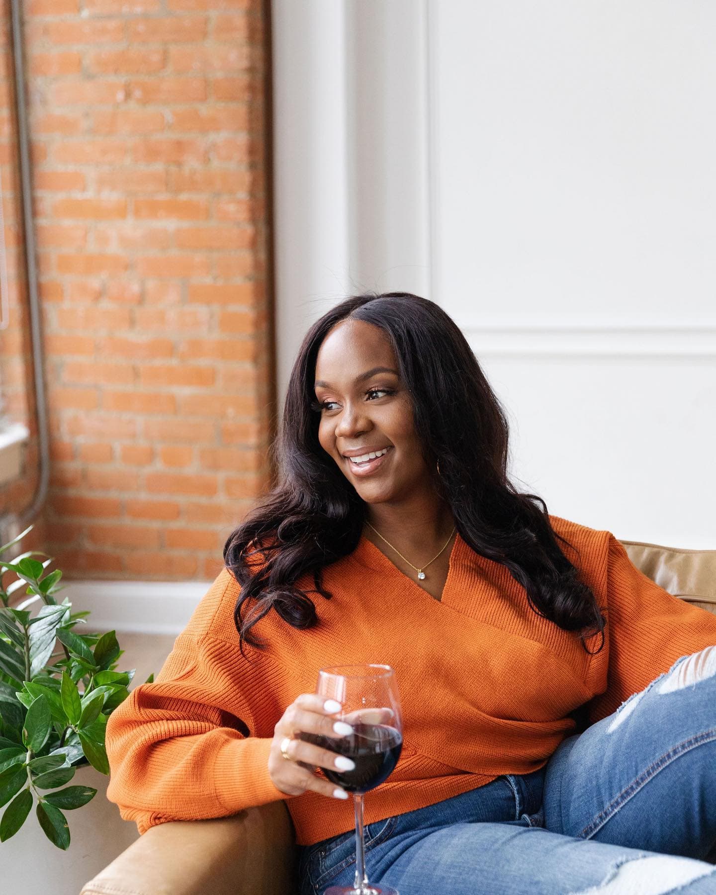 Neké Akua in an orange wrap sweater, holding a glass of wine beside an exposed brick wall.