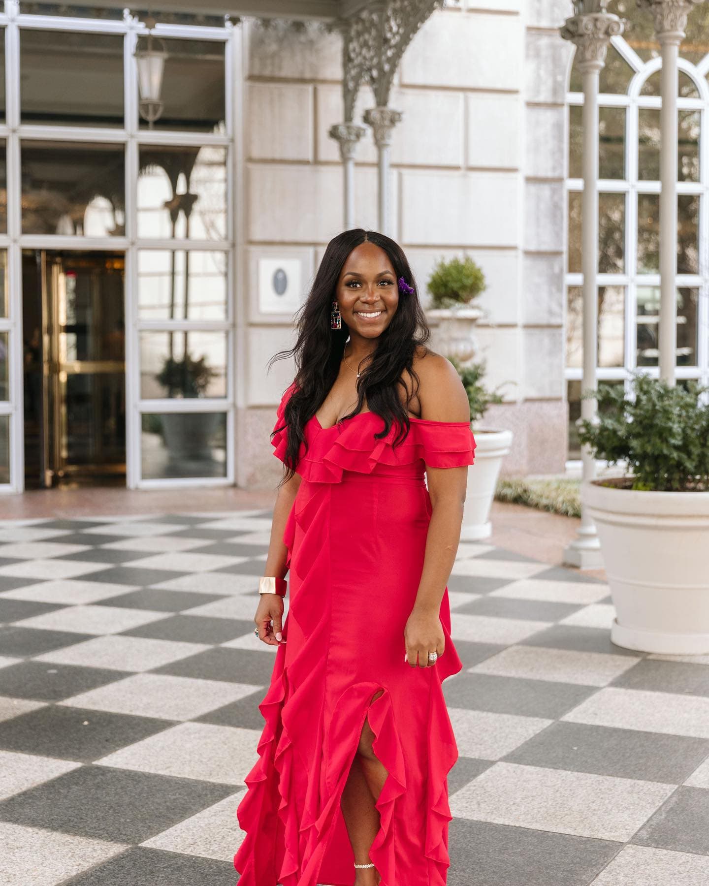 Neké Akua in a red ruffled off-the-shoulder gown on the checkerboard tiled forecourt of a grand white hotel entrance.