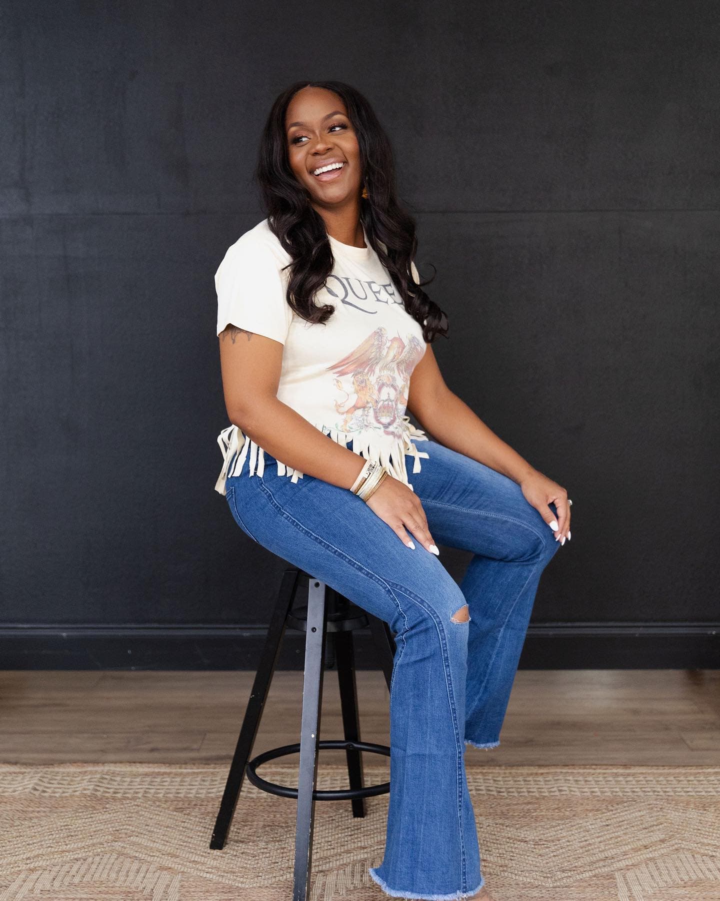 Neké Akua laughing on a stool in a cream graphic tee and flared denim.
