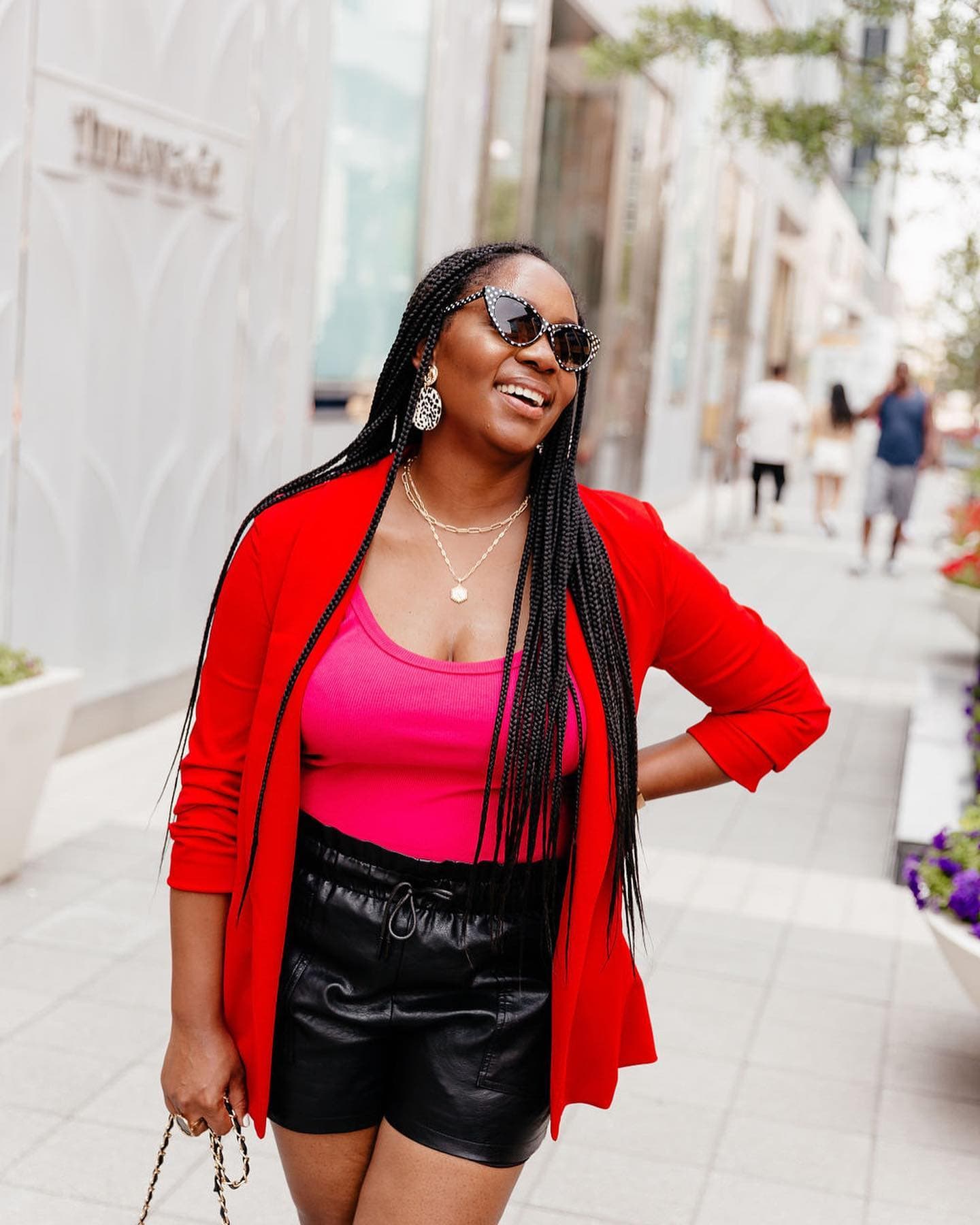 Neké Akua laughing in a red blazer and sunglasses on a city sidewalk.