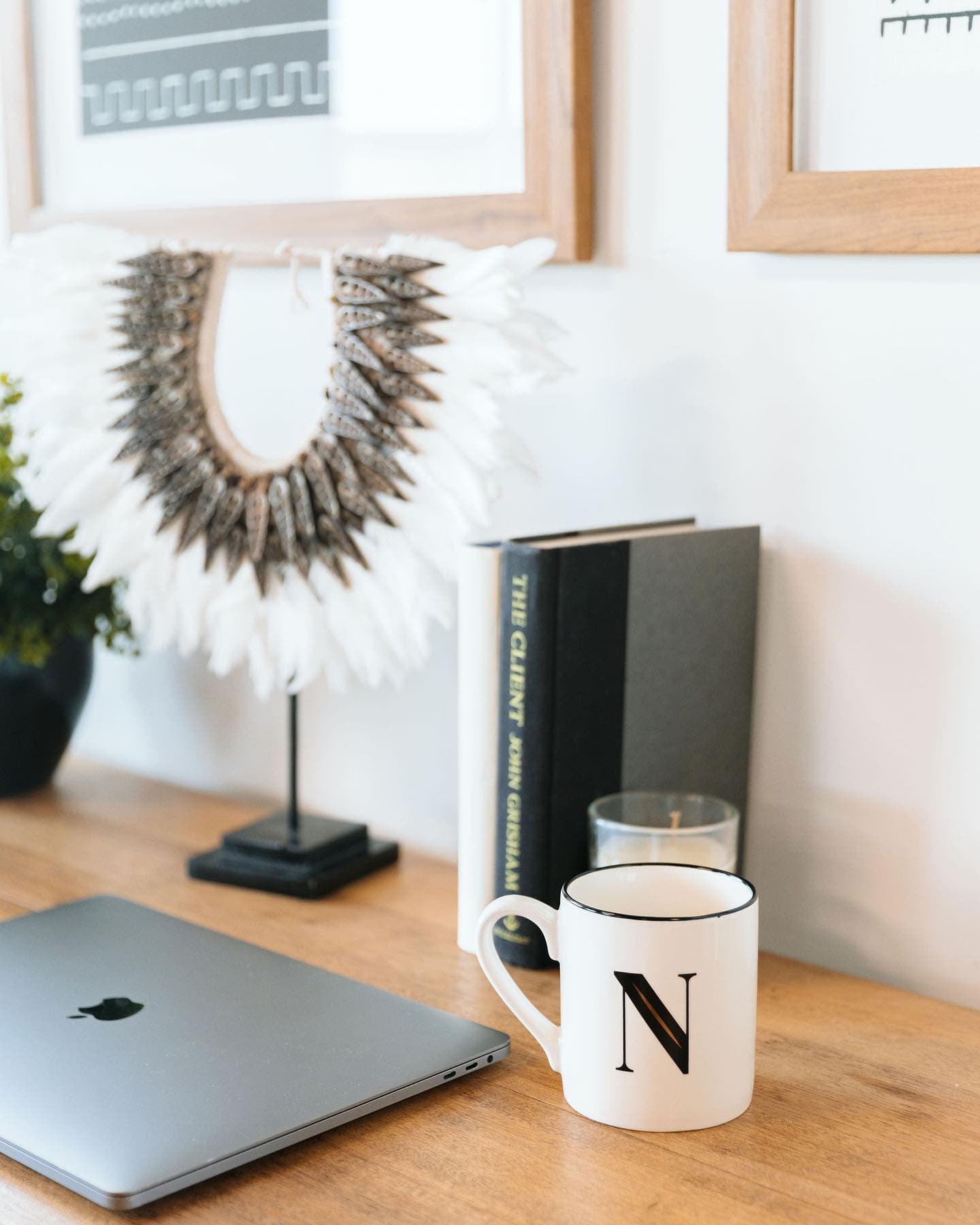 A wooden desk with a laptop, a monogrammed mug, a candle and a feather wall hanging.