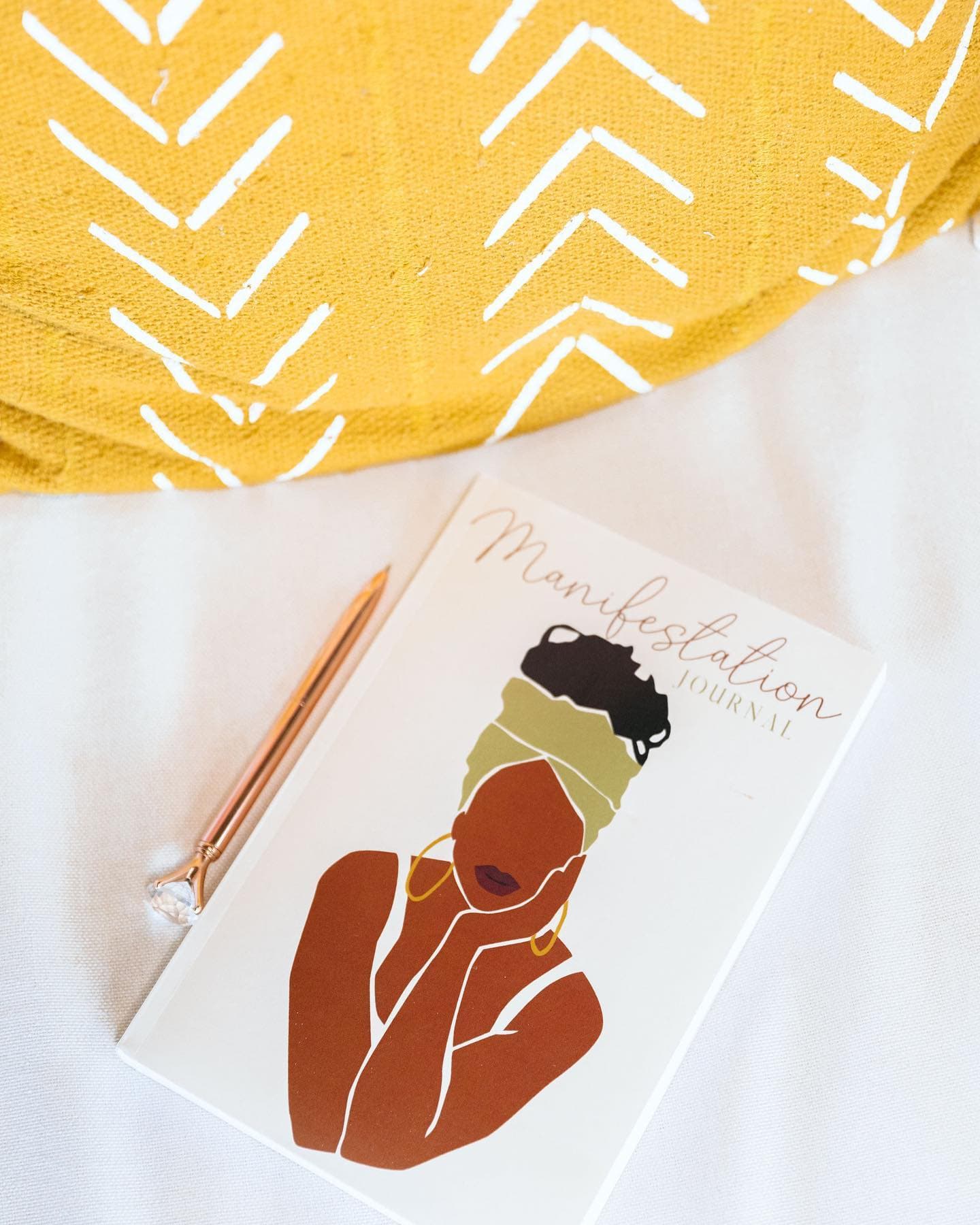 A rose-gold pen resting on a Manifestation Journal against a mustard chevron pillow.
