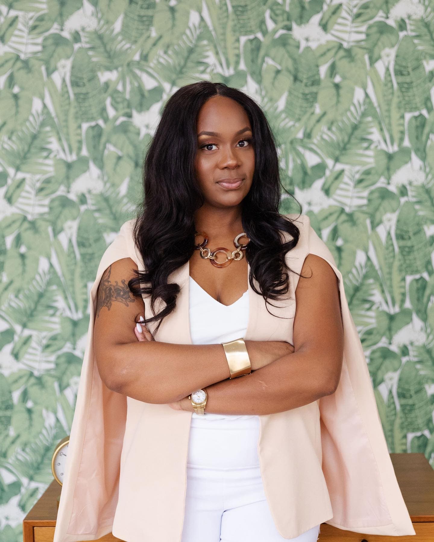Neké Akua with arms folded in a blush cape blazer and white trousers, standing against green palm-print wallpaper.