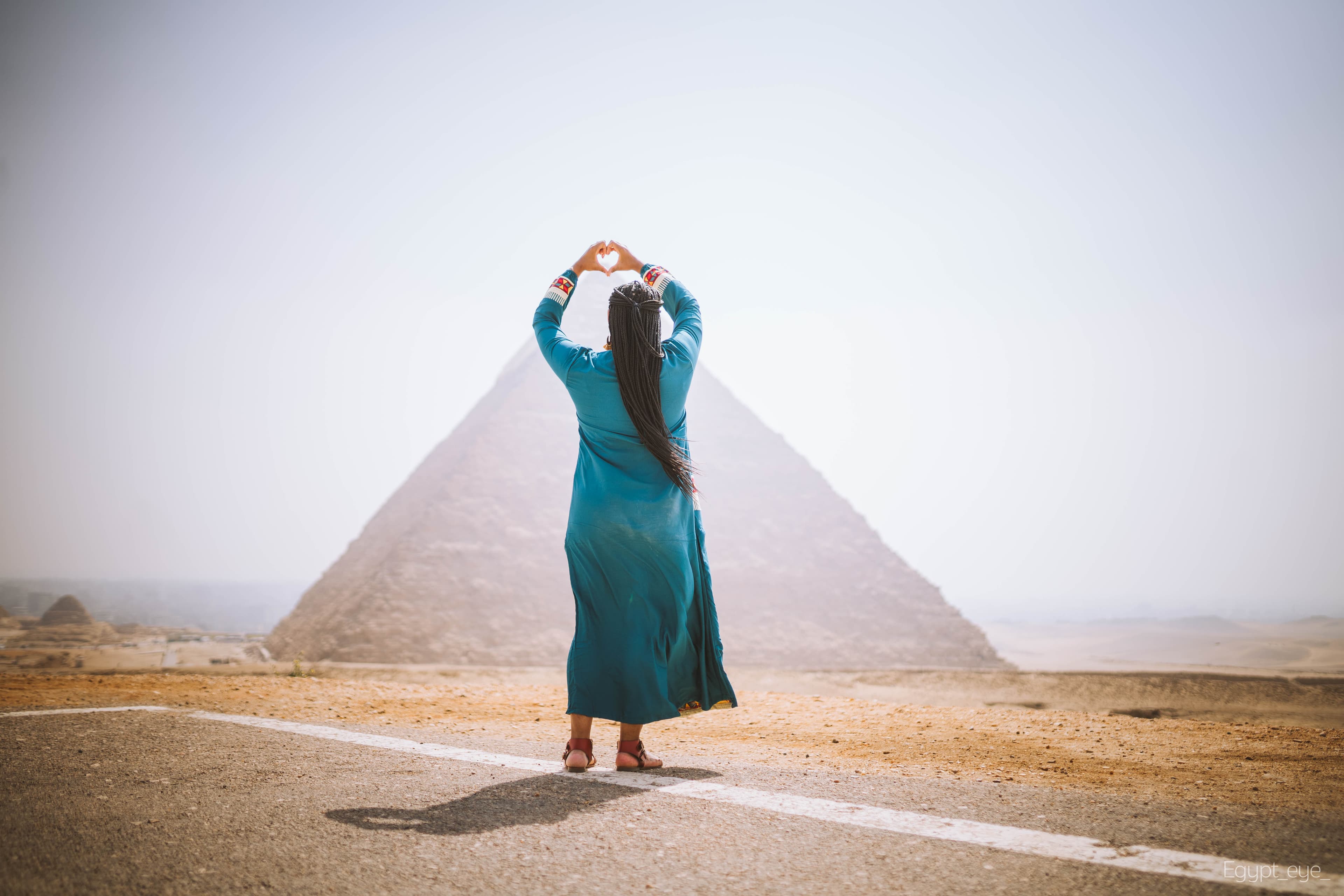 Neké Akua from behind in a teal embroidered kaftan, hands raised in a heart shape before the Great Pyramid of Giza.