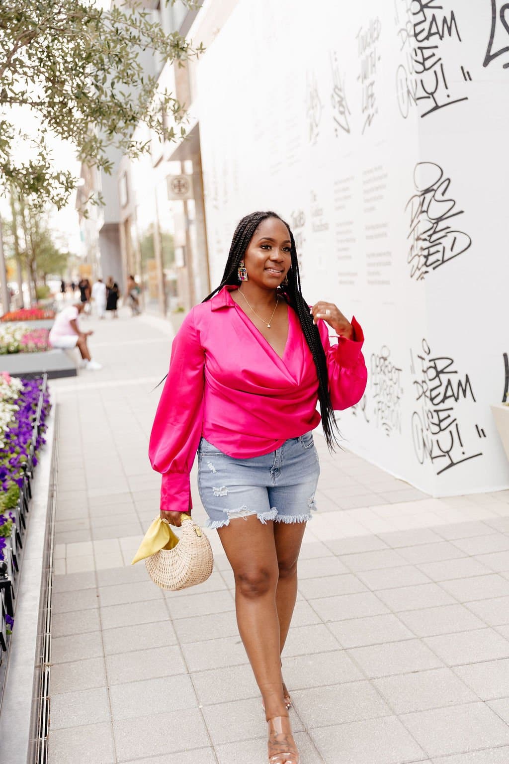 Neké Akua in a hot-pink wrap blouse and denim shorts, glancing back against a graffiti-covered wall, woven bag in hand.