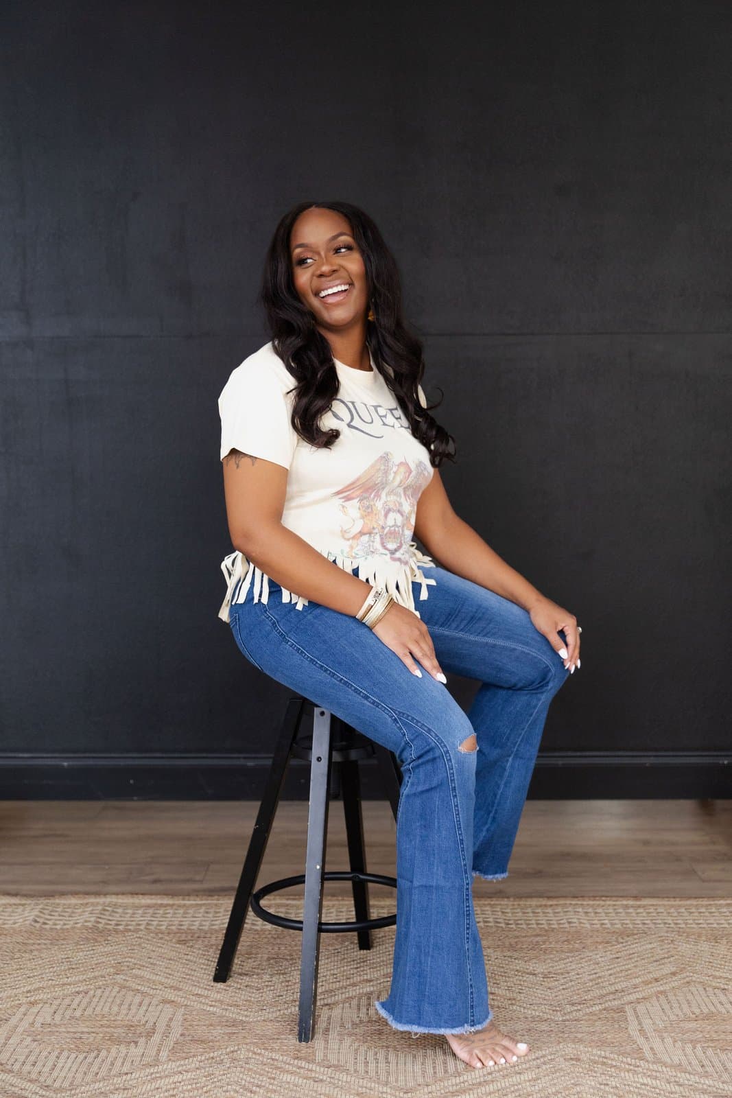 Neké Akua laughing on a stool in a fringed vintage band tee and ripped flare jeans against a charcoal wall.