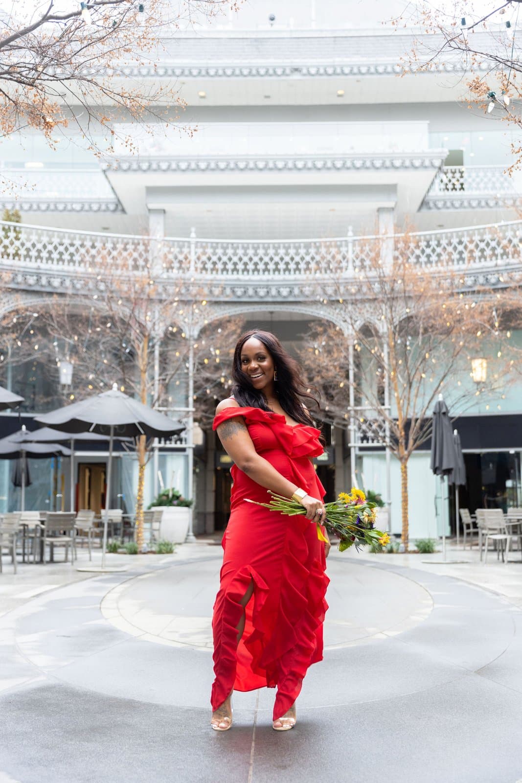 Neké Akua in a red ruffled off-shoulder gown, laughing with a bouquet of wildflowers in a string-lit courtyard.