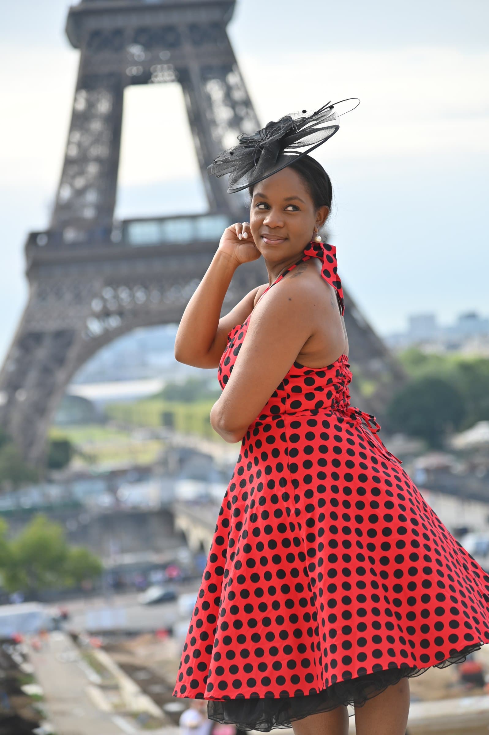 Neké Akua in a red polka-dot dress and black fascinator, glancing back with the Eiffel Tower rising behind her.