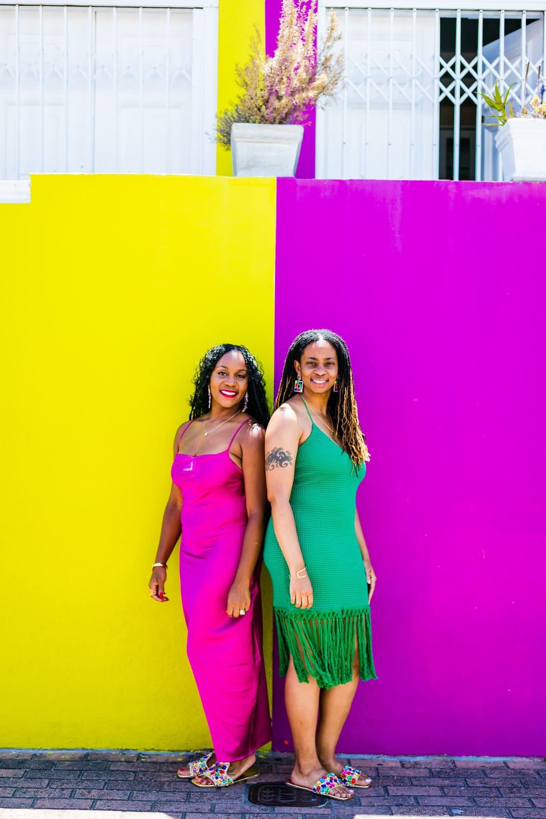 Neké Akua in a fuchsia slip dress and a friend in emerald green, laughing against Bo-Kaap's yellow-and-purple facades in Cape Town.