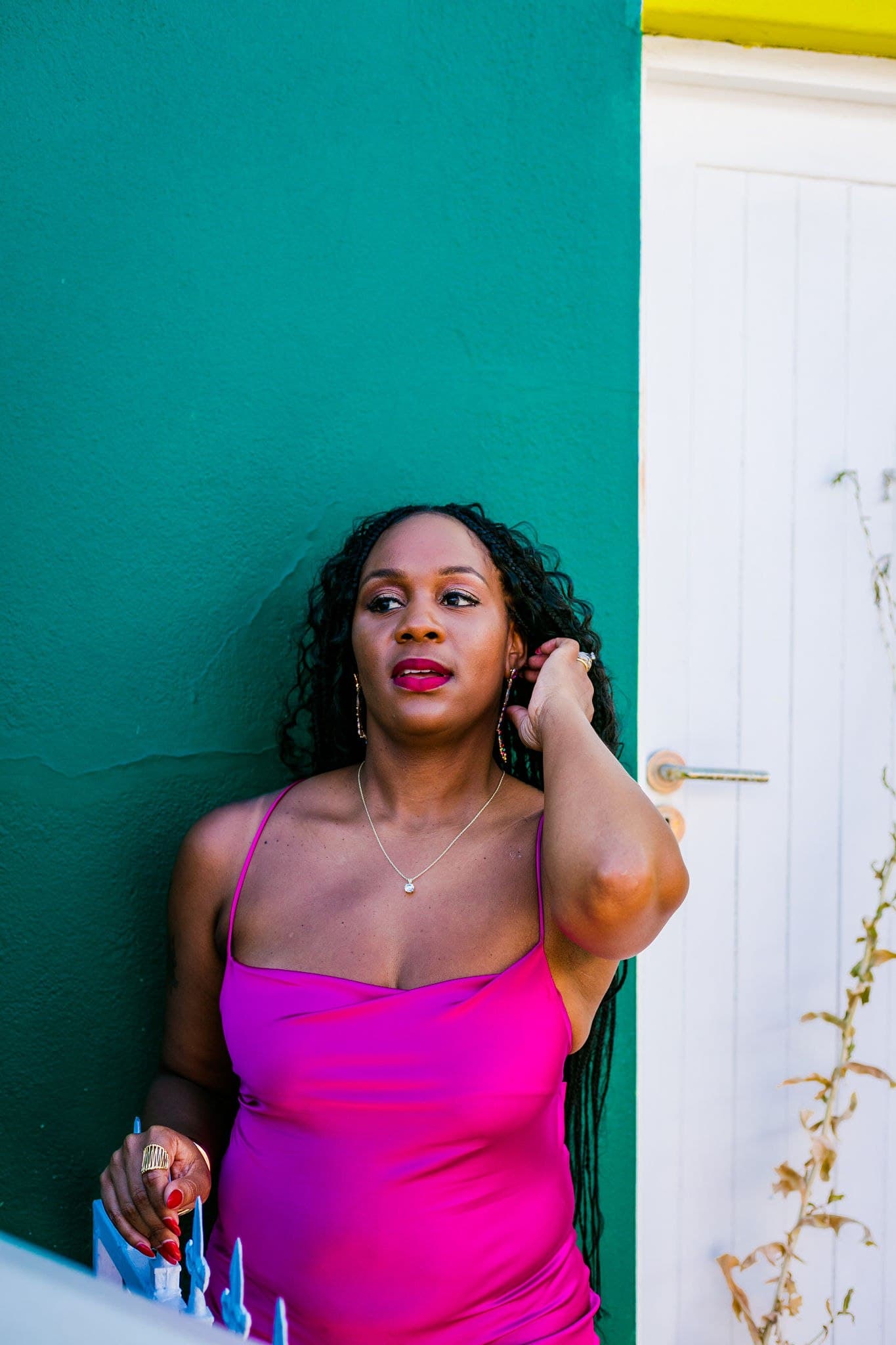 Close portrait of Neké Akua in a fuchsia slip dress against a teal Bo-Kaap wall, tucking back her braids.