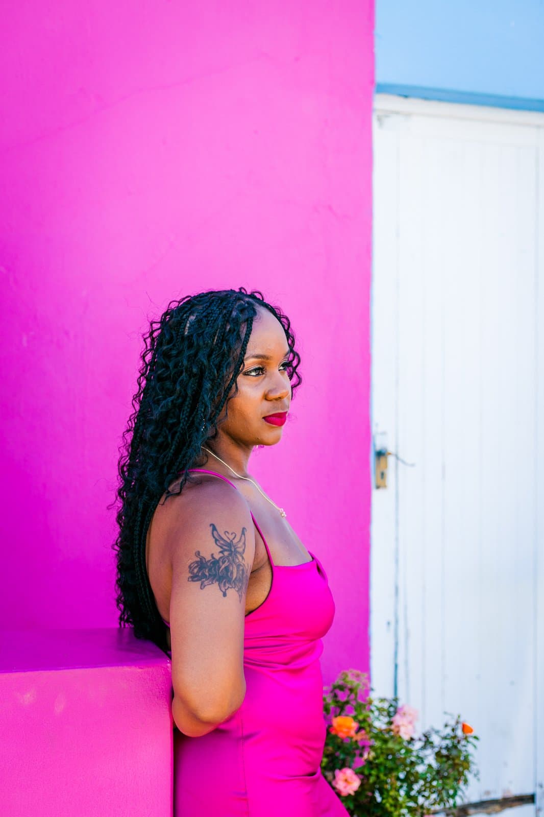 Close profile of Neké Akua in a fuchsia slip dress against a hot-pink Bo-Kaap wall, braids catching the light.
