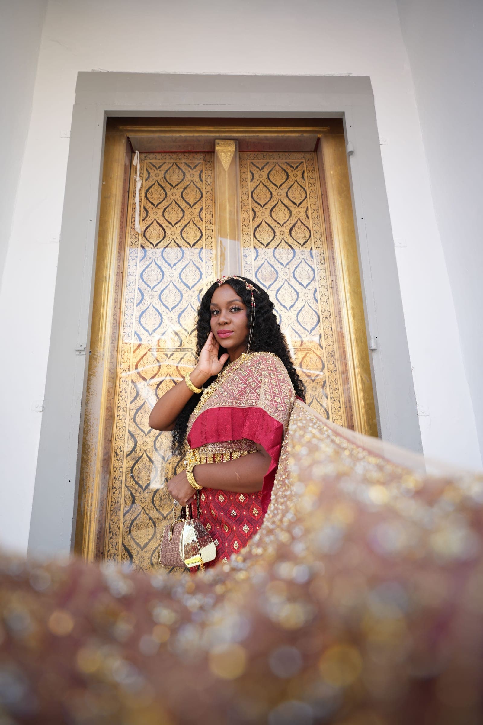 Neké Akua in chut thai framed in a tall gilded doorway, one hand raised to her chin.