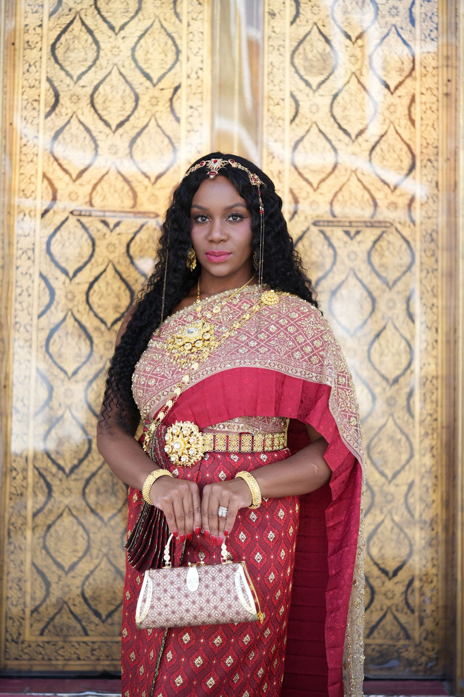 Neké Akua in full chut thai — a red and gold silk sabai with a jeweled belt — standing before a gilded door.