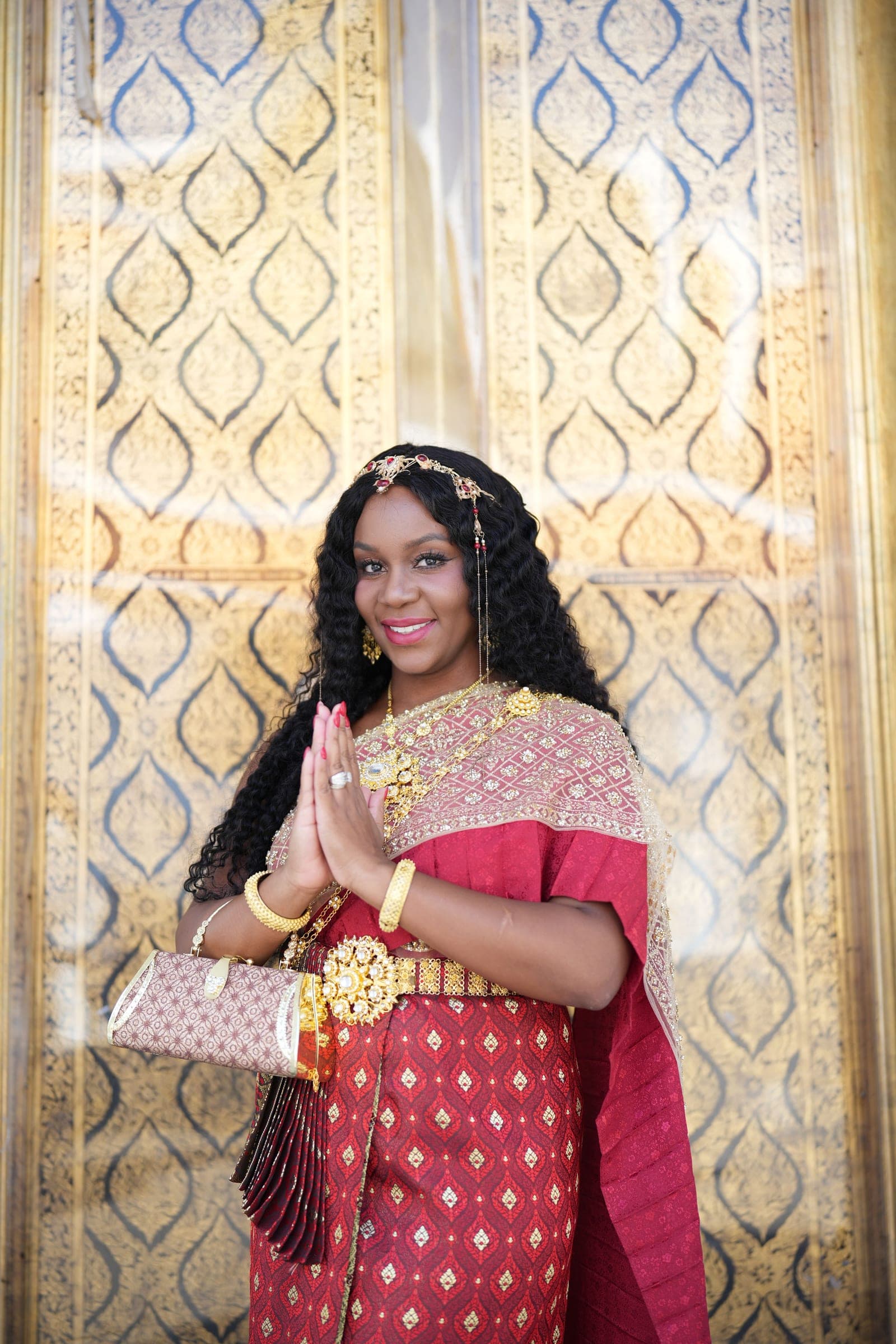 Neké Akua in chut thai with her palms pressed together in a wai, gold-patterned doors behind her.