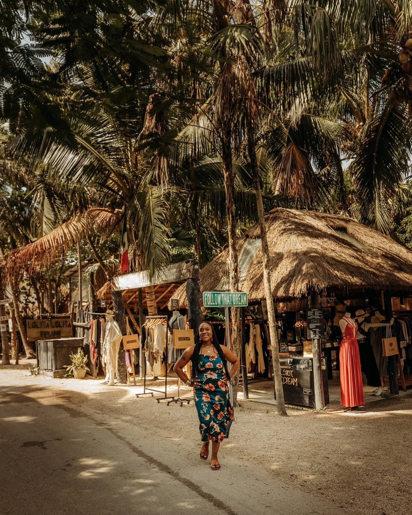 Neké Akua in a floral dress walking past boutique stalls under palm trees on a sandy Tulum street.