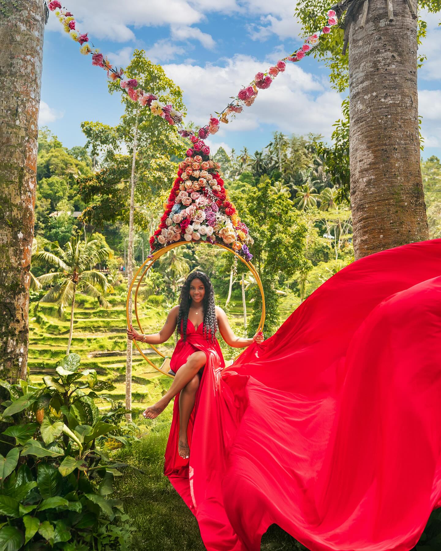 Neké Akua in a red gown, fabric sweeping in the wind, on a flower-hung swing above Balinese rice terraces.