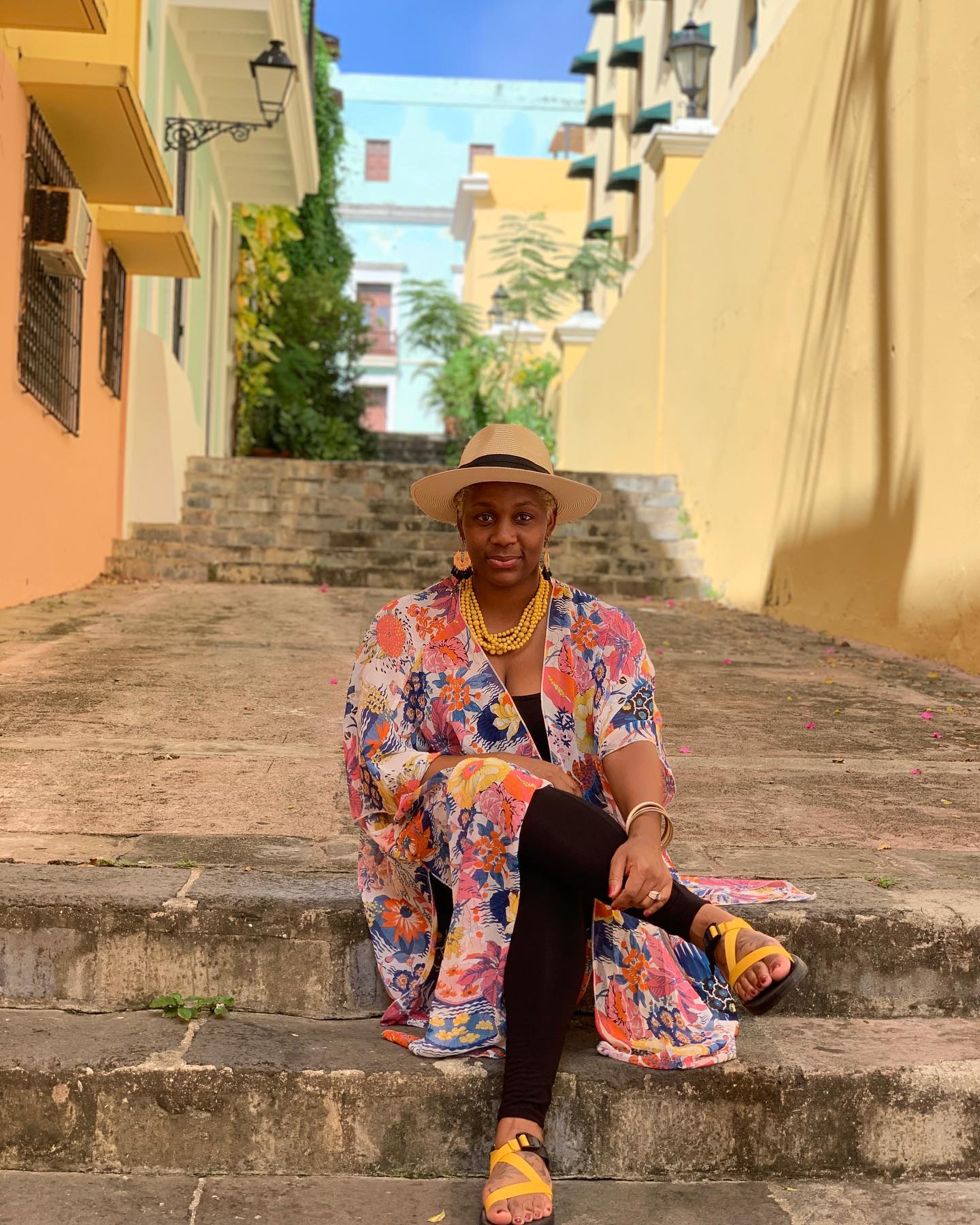 Neké Akua in a floral kimono and sun hat, seated on stone steps between colorful buildings in Old San Juan.