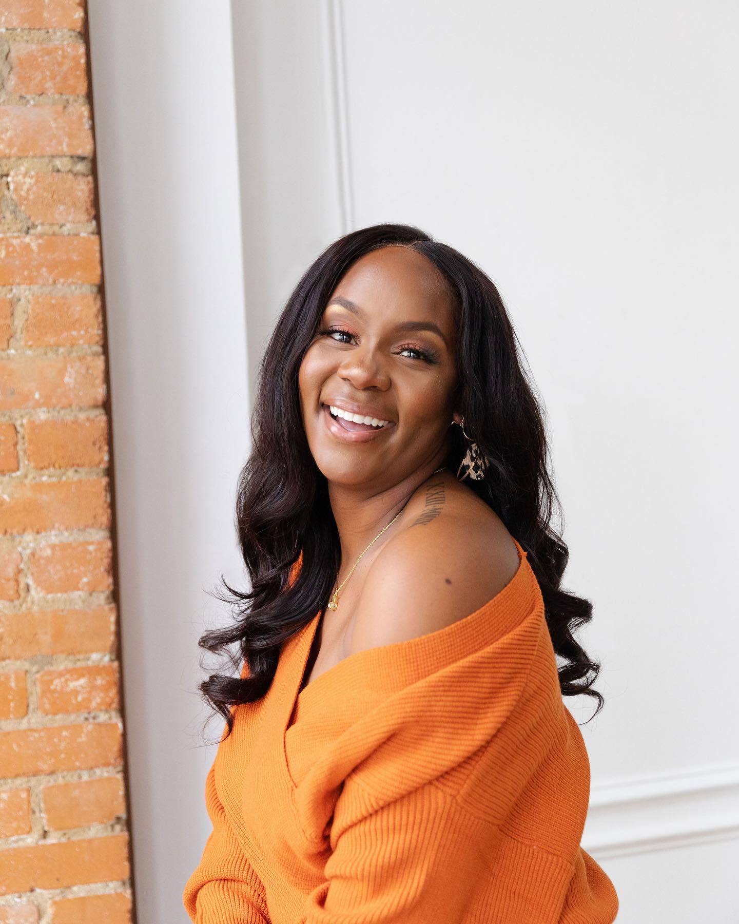 Neké Akua laughing in a burnt-orange sweater against an exposed brick wall.