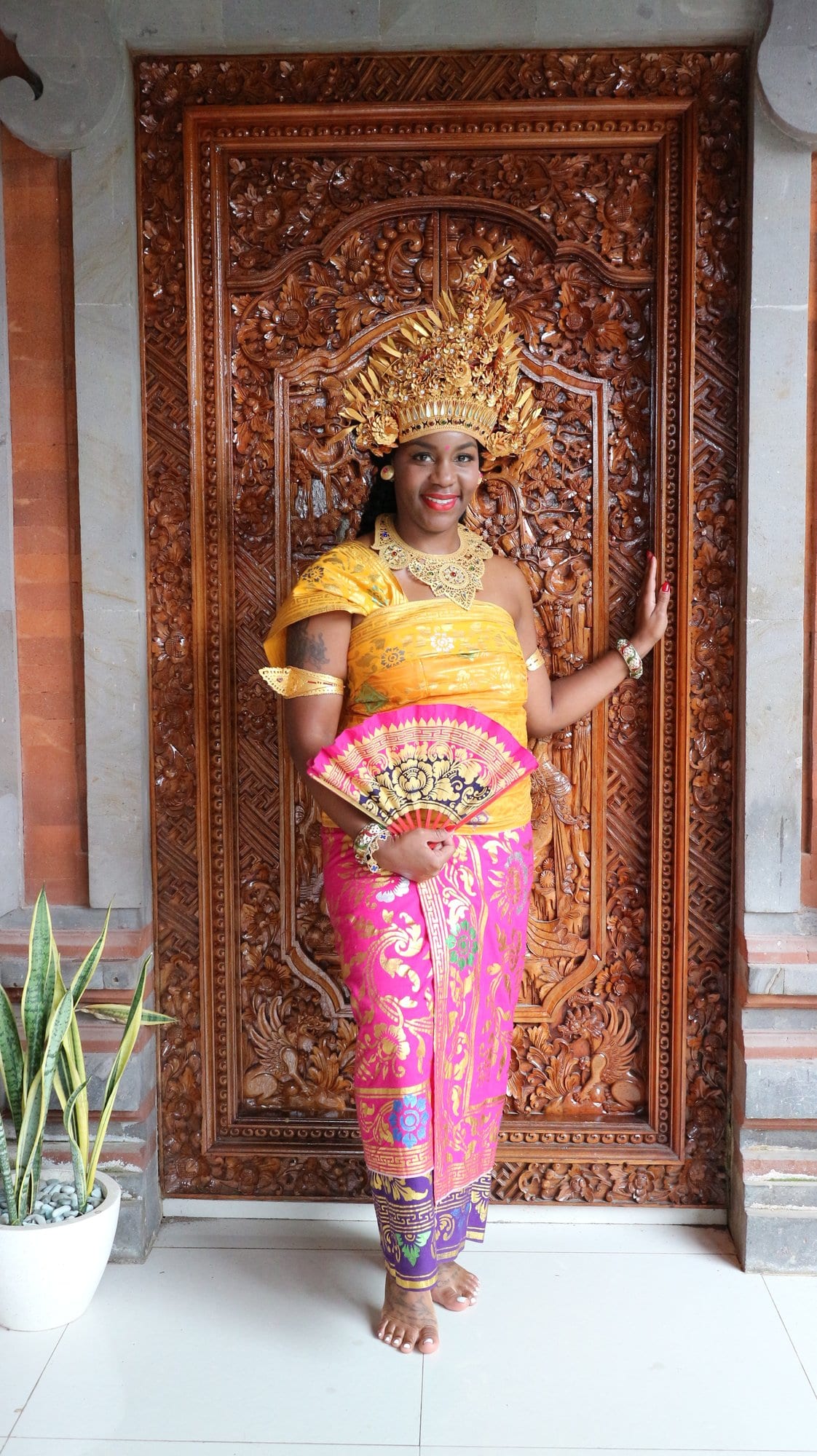Neké Akua in an elaborate gold Balinese headdress and pink-and-gold sarong, fan in hand before a carved wooden door.