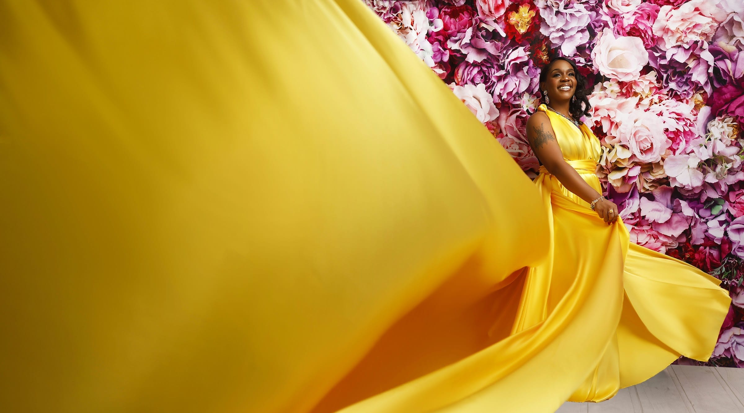 Neké Akua laughing in a gold satin gown against a wall of blush and burgundy flowers, the train of the dress caught mid-motion across the frame.