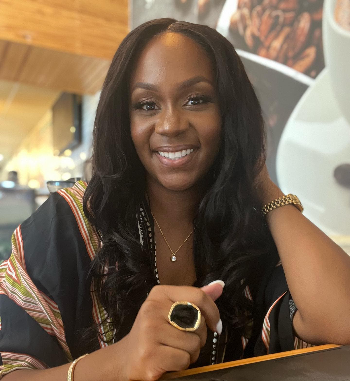 Neké Akua laughing at a coffee-shop table in a striped black kimono, gold watch and statement ring catching the light.