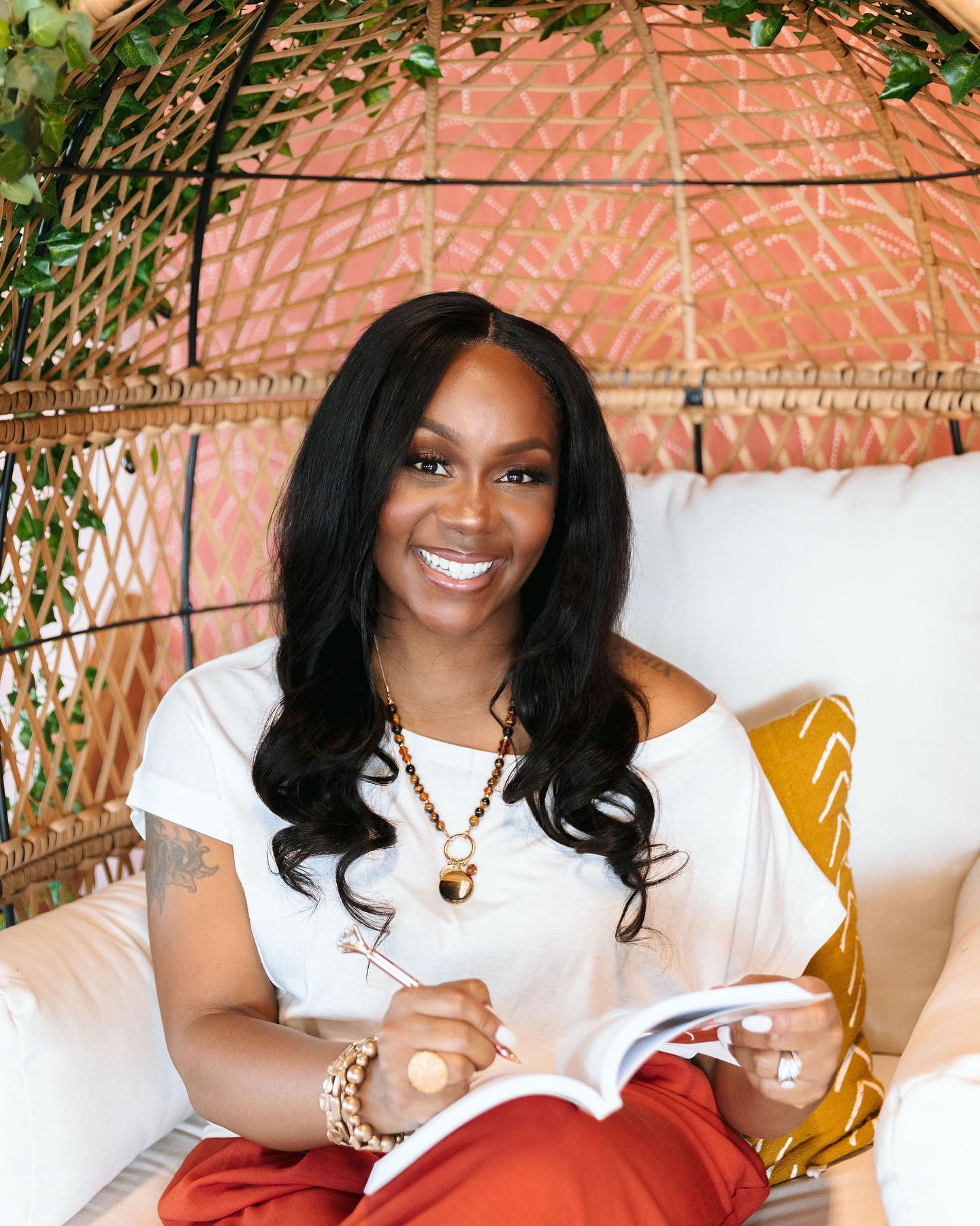 Neké Akua journaling inside a wicker egg chair against a coral wall trimmed with greenery.