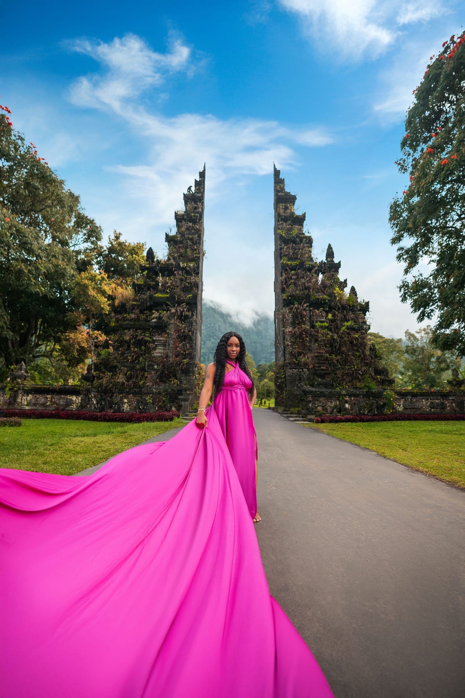 Neké Akua at Bali's Handara Gate in a magenta gown, its train sweeping the full width of the road behind her.