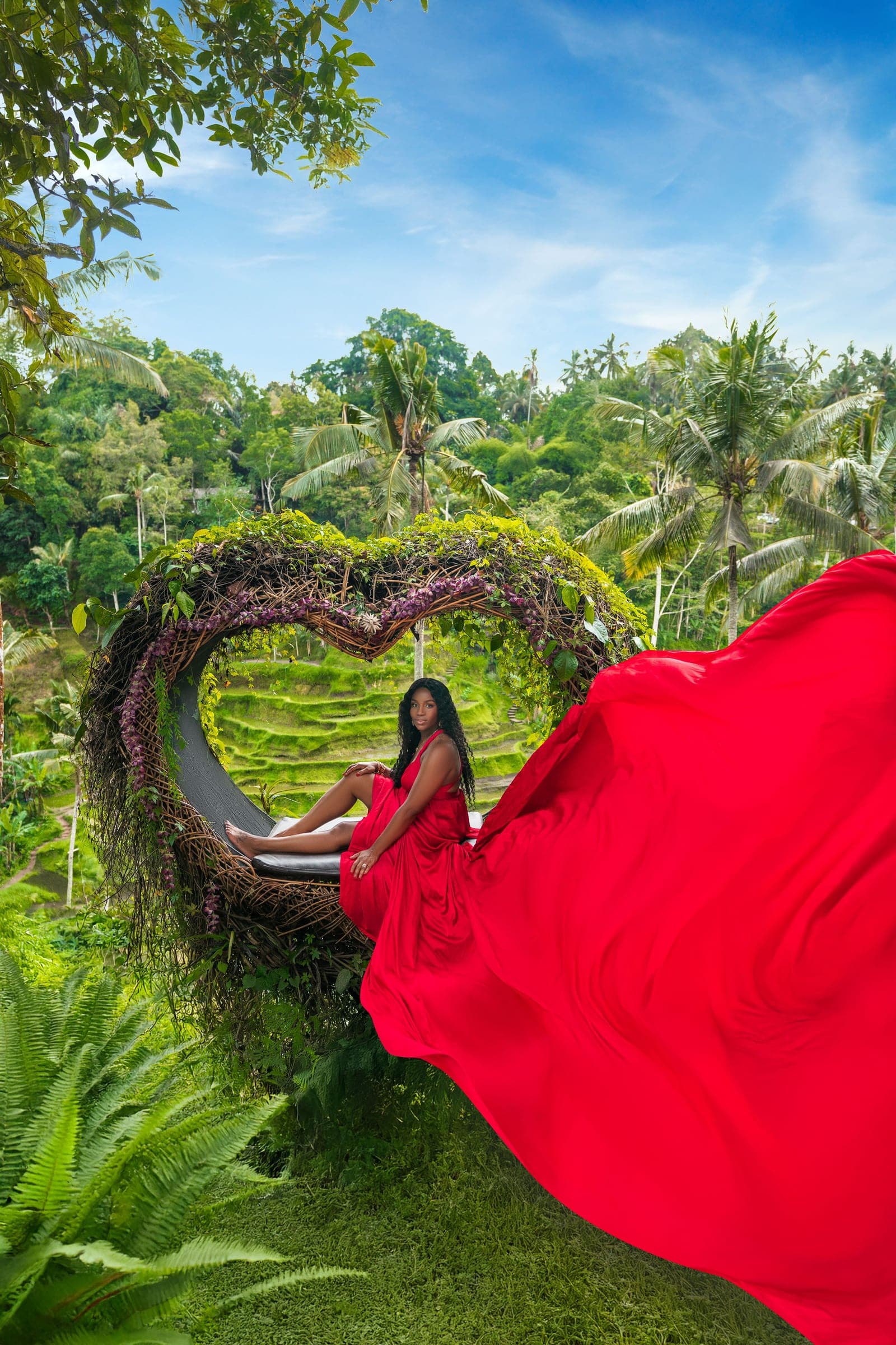 Neké Akua in a red gown inside a heart-shaped woven nest, jungle behind her and the dress's train sweeping out across the grass.