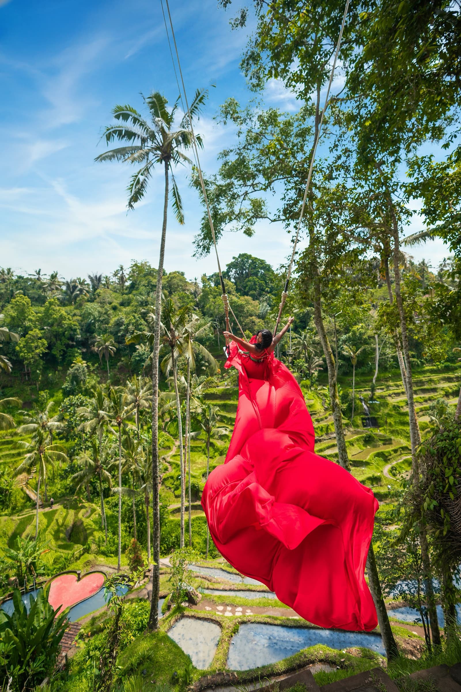 Neké Akua in a red gown on a flower-hung jungle swing in Bali, the train of the dress flying out over the treetops.