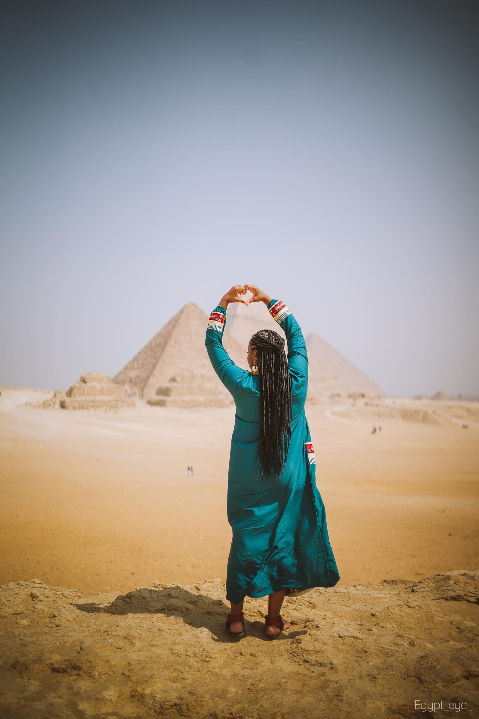 Neké Akua from behind with both arms raised over her head, framed against a pyramid under a pale, hazy sky.