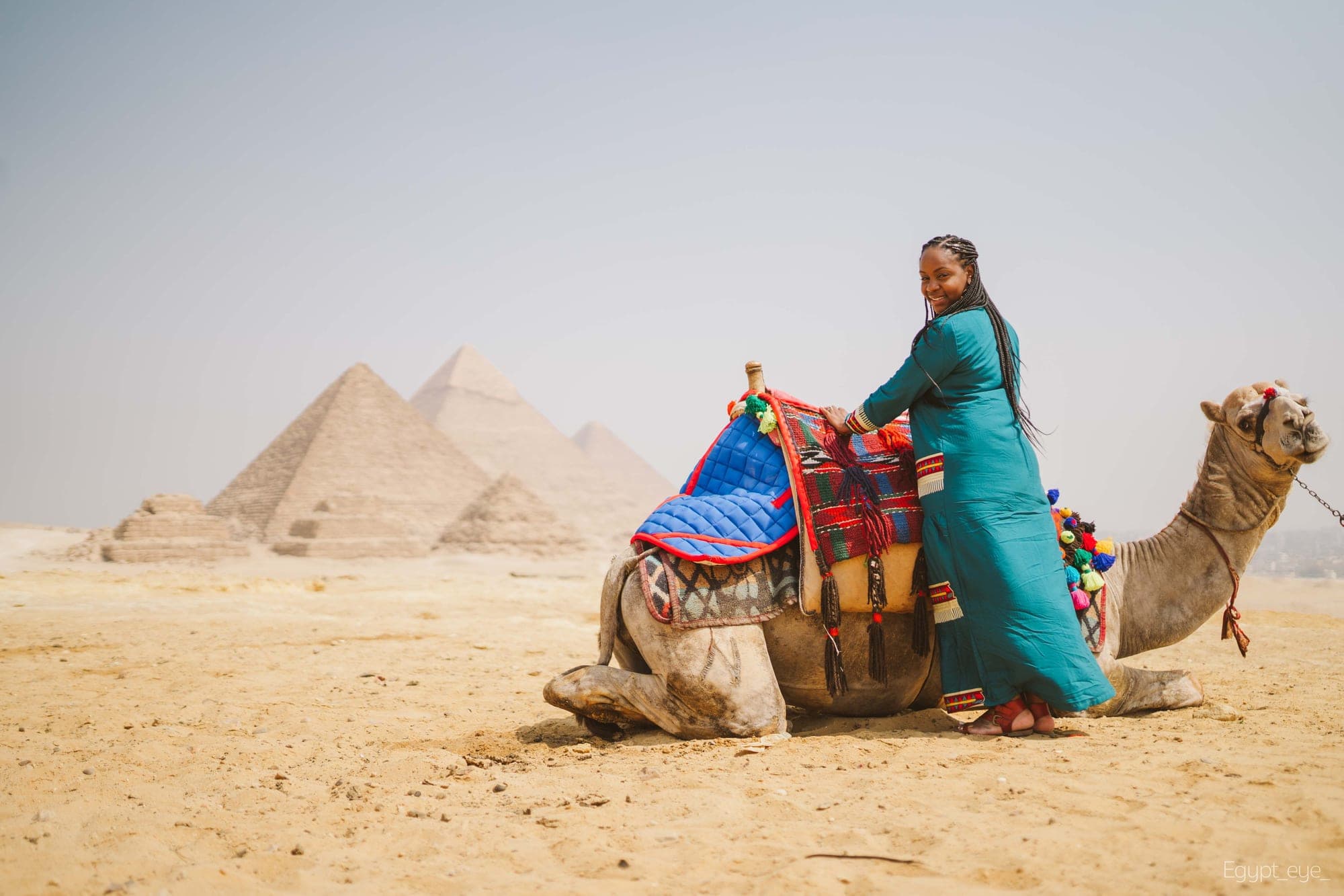 Neké Akua standing beside a resting camel in a red and blue tasselled blanket, three pyramids on the horizon behind them.