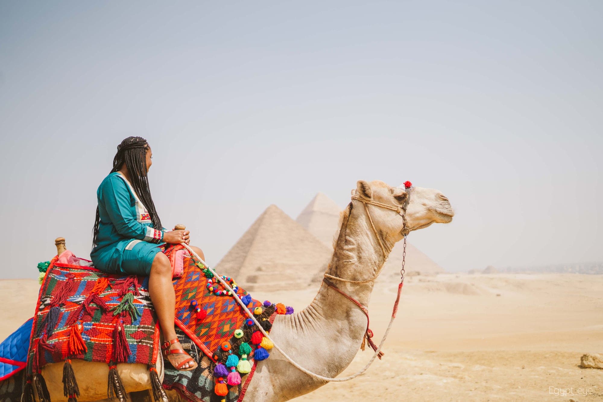 Neké Akua seated on a decorated camel in a teal kaftan, a pyramid standing in the haze behind them.