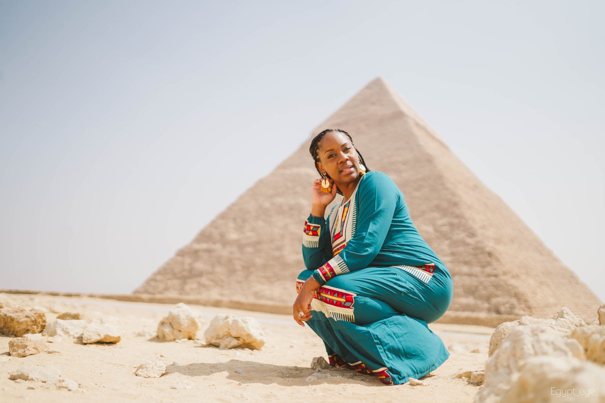 Neké Akua crouching on the sand in a teal embroidered kaftan, the weathered blocks of the Great Pyramid filling the frame behind her.