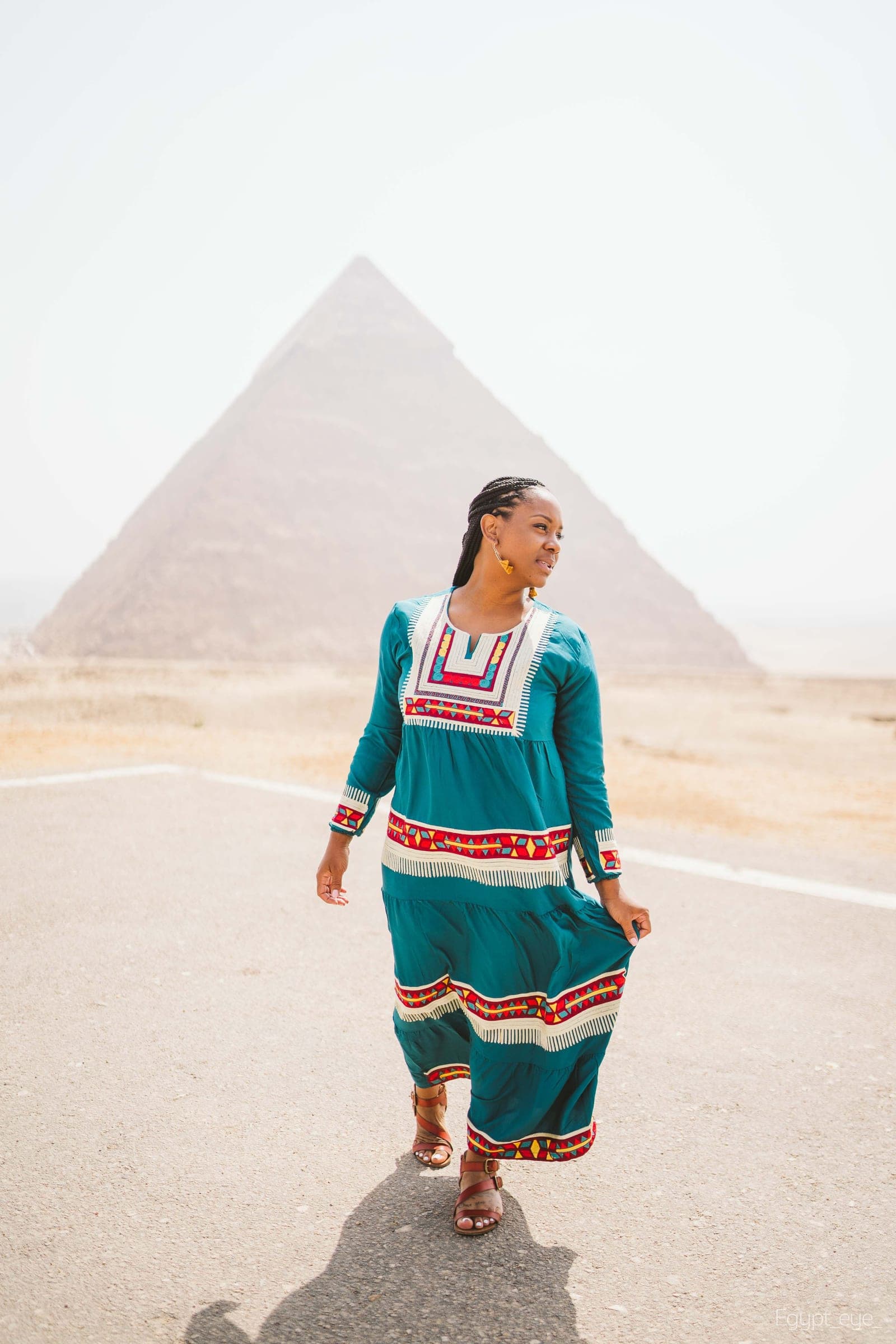 Neké Akua walking up the middle of the desert road at Giza in a teal kaftan, a pyramid rising dead center ahead of her.